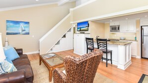A spacious living area with a brown leather sofa, a glass coffee table, and a woven chair. A staircase leads to a kitchen with white cabinets, a stainless steel refrigerator, and a built-in island with bar stools.