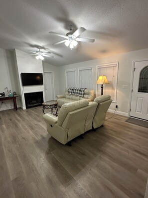 A living room with a beige leather recliner, a ceiling fan, a flat-screen TV, and a fireplace.