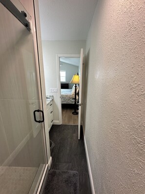 A narrow hallway with a shower area on the left, a bedroom with a bed and a lamp on the right, and a door leading to another room.