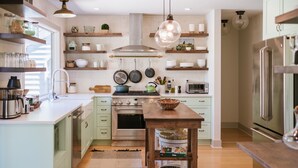 A modern kitchen with white cabinets, a stainless steel stove, and hanging pots. There are wooden shelves above the stove and a wooden table in the center. - Luxe remodel, three King ensuites, walk to 3rd St (McMinnville)