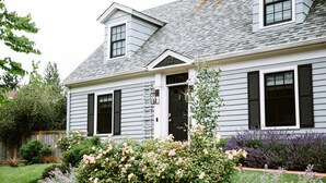 A two-story house with a dark front door, a small porch, and a well-maintained garden with various flowers and shrubs. - Luxe remodel, three King ensuites, walk to 3rd St (McMinnville)