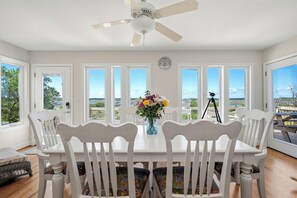 Un comedor con mesa y sillas blancas, ventilador de techo, reloj y vistas amplias a través de grandes ventanas.
