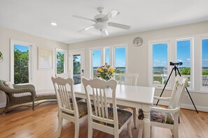 A dining area with a white table, chairs, a ceiling fan, and large windows with a view. - Sound Bite: Ocean View Home with Dock on Eastern Channel! (Oak Island)