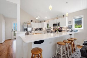 Une cuisine moderne avec des meubles blancs, un îlot central doté de tabourets, un ventilateur de plafond et des luminaires suspendus.