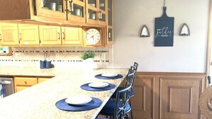 Een keuken met houten kastjes, een granieten aanrecht en een klok. Er staan barkrukken bij en er hangt een lamp aan de muur.