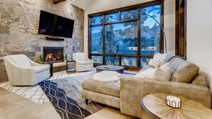 A spacious living room with a stone wall, a fireplace, a large window with a view of trees, and a modern TV mounted on the wall. - 🏆 Breck Ski Views | Gondola Access | Hot Tub | 3 Kings | Sleeps 8 (Breckenridge)