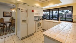 Eine Küche mit weißen Schränken, einem Kühlschrank, einem Backofen und einem Fenster mit Blick auf Palmen.