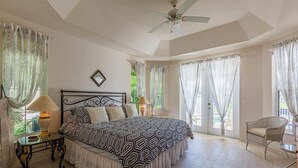A spacious bedroom with a large bed, a ceiling fan, and a view of the outdoors through large windows.