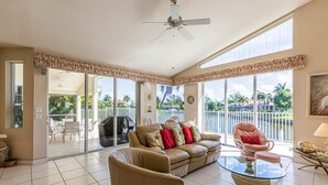 A spacious living room with a beige sofa, a glass coffee table, and a chair. There is a balcony with a view of palm trees and a pool.