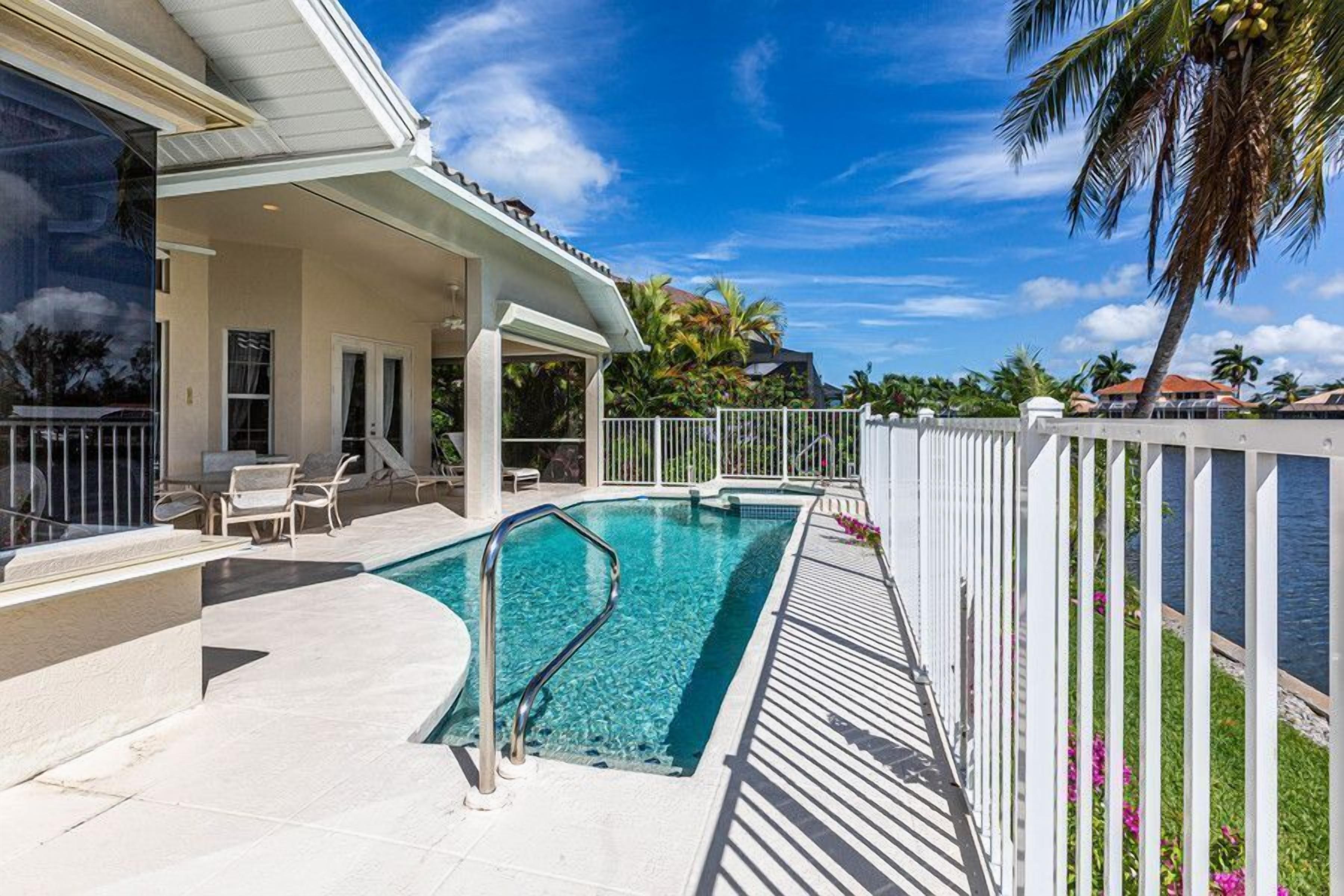 A swimming pool with a white fence, lounge chairs, and a covered area with a view of the ocean.