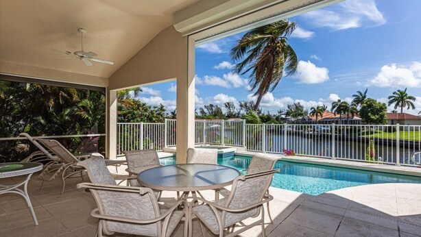 A poolside area with a round table and chairs, a ceiling fan, and a view of a body of water with palm trees.