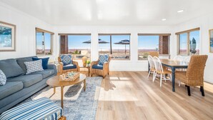 A spacious living room with a grey sofa, wicker chairs, a wooden dining table, and large windows offering a view of the beach. - 2 Bed Oceanfront w/ patio on the Sand in Quiet West Newport (Newport Beach)