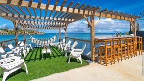 Een houten pergola met uitzicht op de oceaan, witte stoelen en een tafel met drankjes.