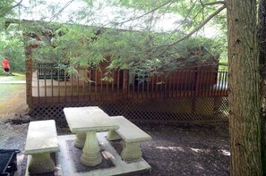 A wooden deck with a lattice fence, surrounded by trees. There are two stone picnic tables in the foreground. -  2 bedroom 1 bath cabin      (Oneida)