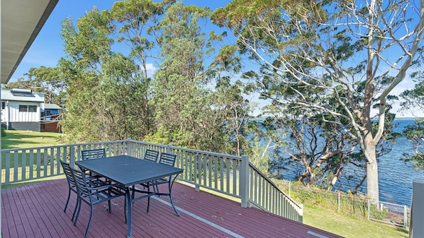 A deck with a table and chairs overlooking a body of water, surrounded by trees and a building. - Palm Beach Waterfront Retreat (Sanctuary Point)