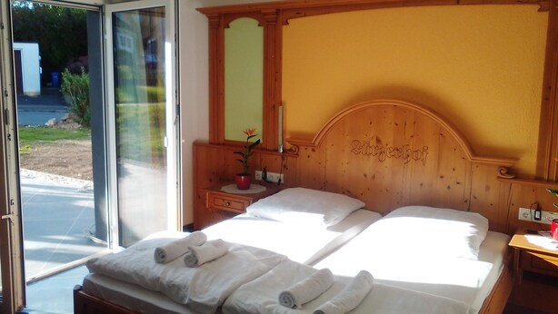 A hotel room with a wooden headboard, a bed with white linens, a nightstand with a lamp, and a sliding glass door leading to an outdoor area.