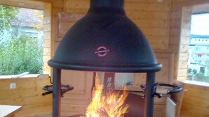 A wood-burning stove with a glass door, set in a wooden cabin with a window and a table.