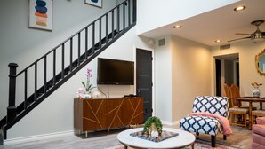 Een moderne woonkamer met een trap, een ronde salontafel, een fauteuil met een patroon en een eethoek met een spiegel en tafel.