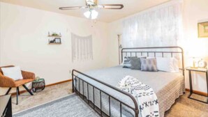 Un dormitorio con cama, una silla, una mesita de noche y un ventilador de techo, además de una ventana con cortinas.
