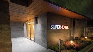 Exterior de un hotel de diseño moderno, con un llamativo letrero 'SuperHotel', una entrada bien iluminada y un área ajardinada con plantas y un hidrante.