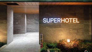 Exterior de un hotel llamado "Superhotel" con un diseño moderno, que presenta un letrero iluminado llamativo, fachada de piedra y un área de entrada bien iluminada.