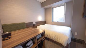 A compact hotel room with a single bed, a small desk with a chair, and a window providing natural light.