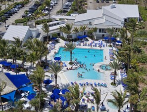 An aerial view of a resort with a large central pool surrounded by lounge chairs and umbrellas, palm trees, and a parking lot.