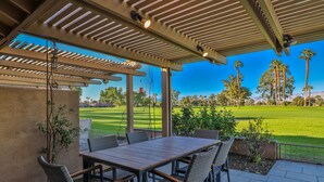 Eine Terrasse im Freien mit Tisch und Stühlen, einer Pergola und Blick auf ein grünes Feld und Bäume.