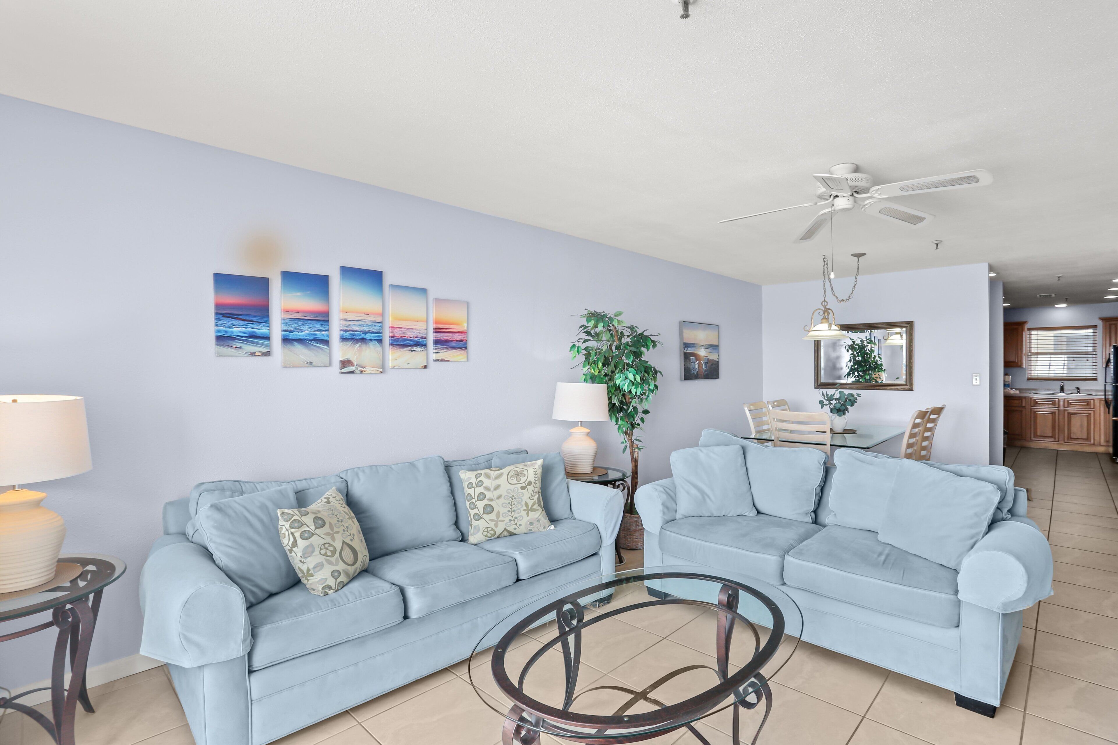 A living room with two light blue sofas, a glass coffee table, a ceiling fan, and wall art depicting sunsets.