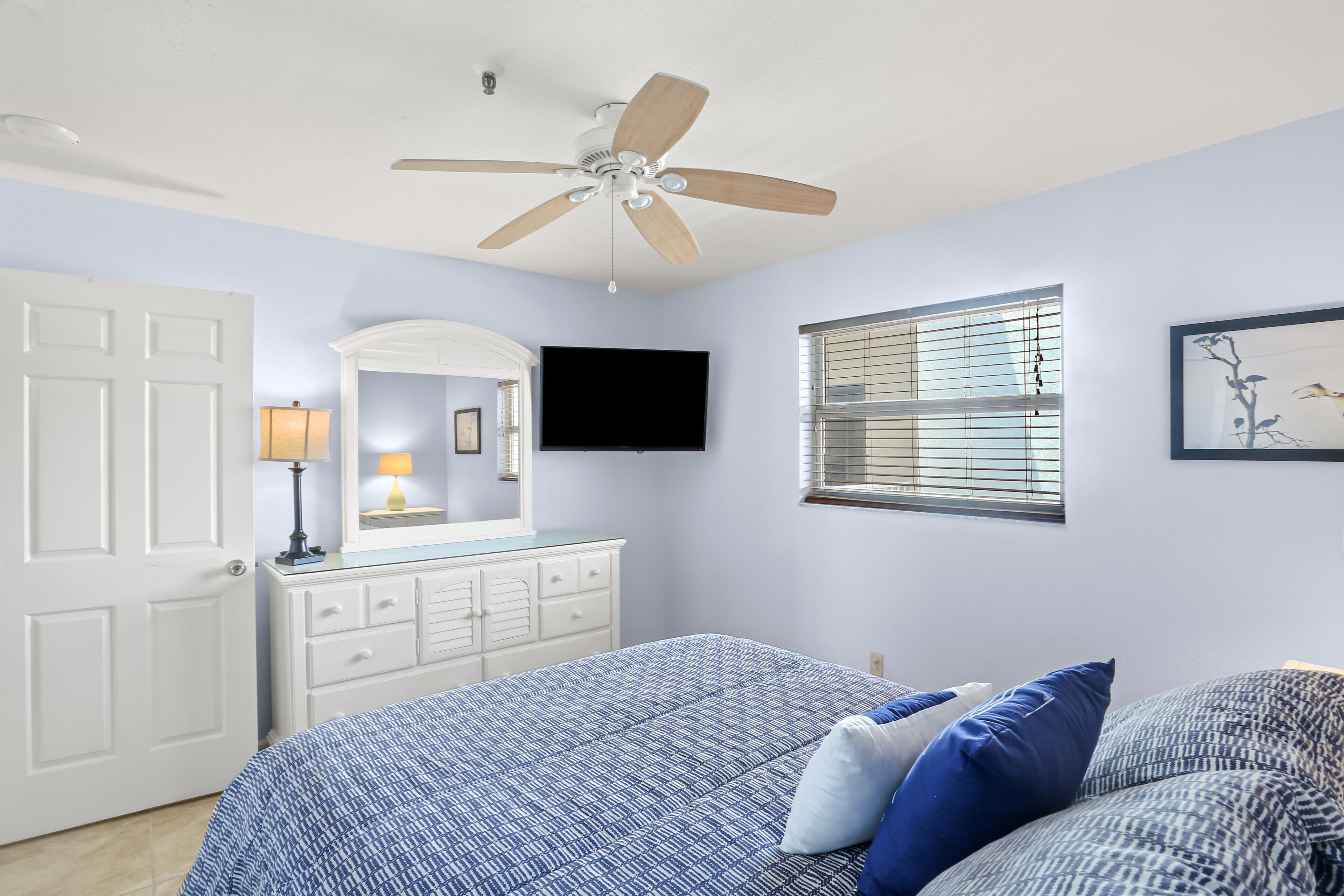 A bedroom with a bed, a ceiling fan, a dresser, a TV, and a window with blinds.