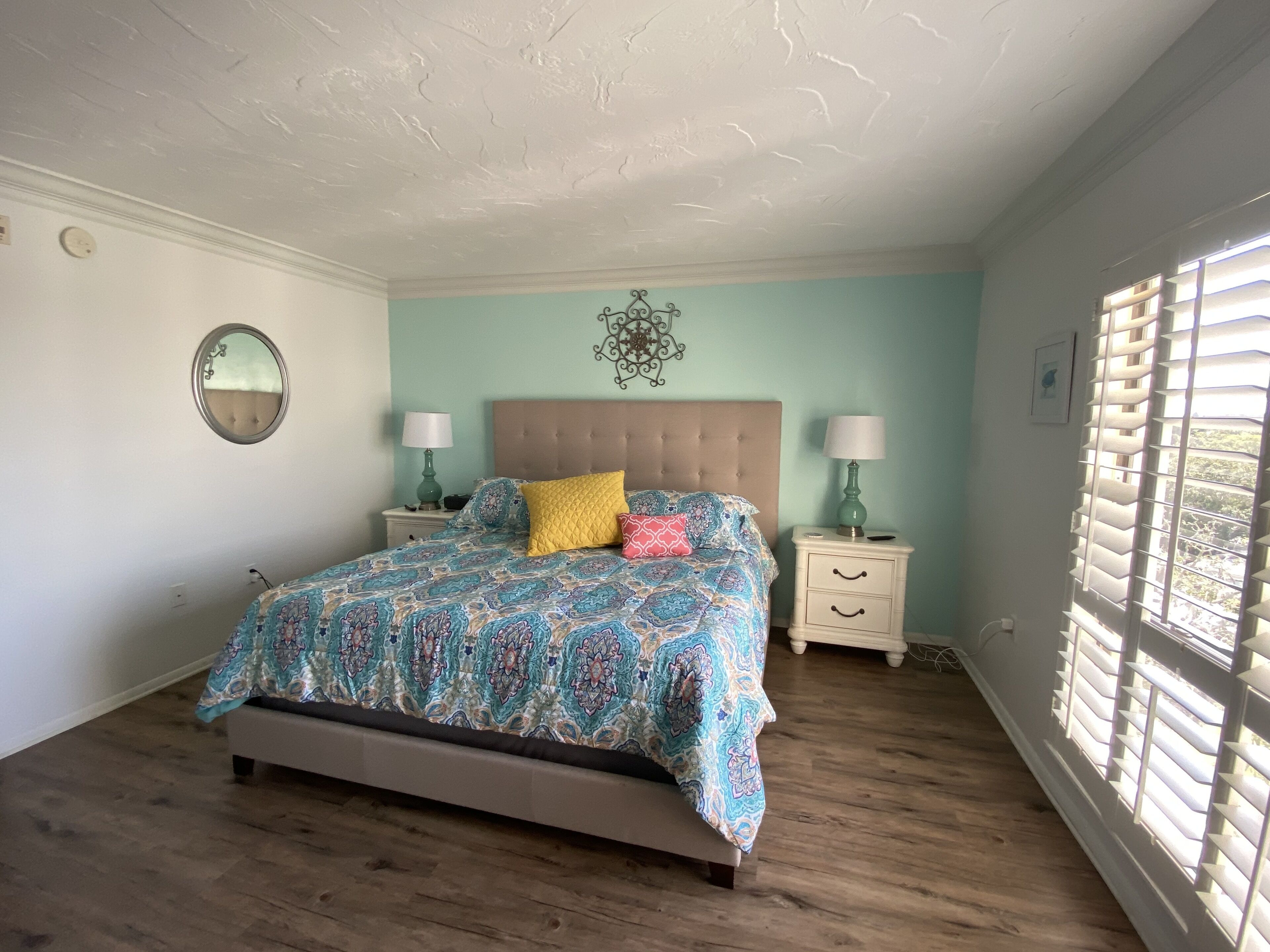 Een slaapkamer met een bed, twee nachtkastjes, een spiegel en een raam met jaloezieën.