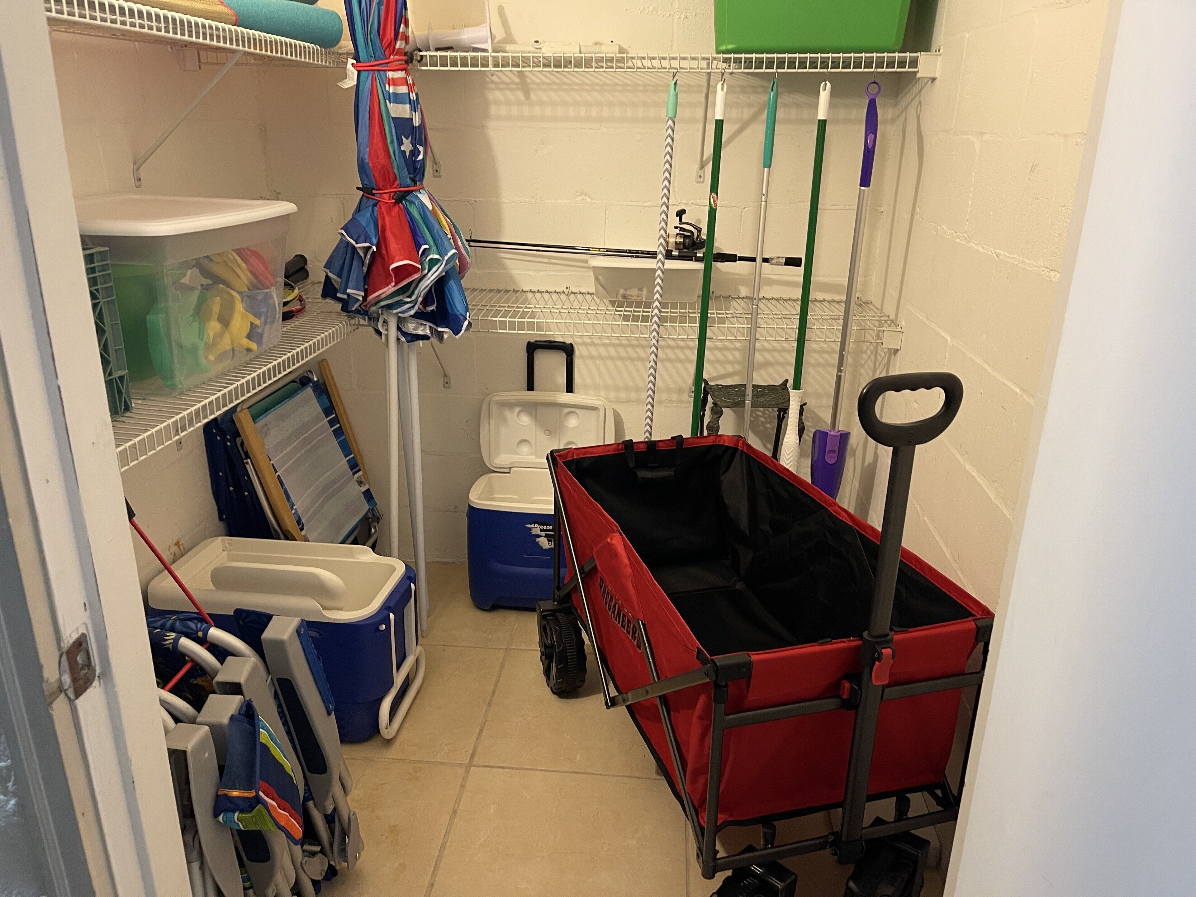 A small utility room with a red cart, blue cooler, and various cleaning supplies on shelves.