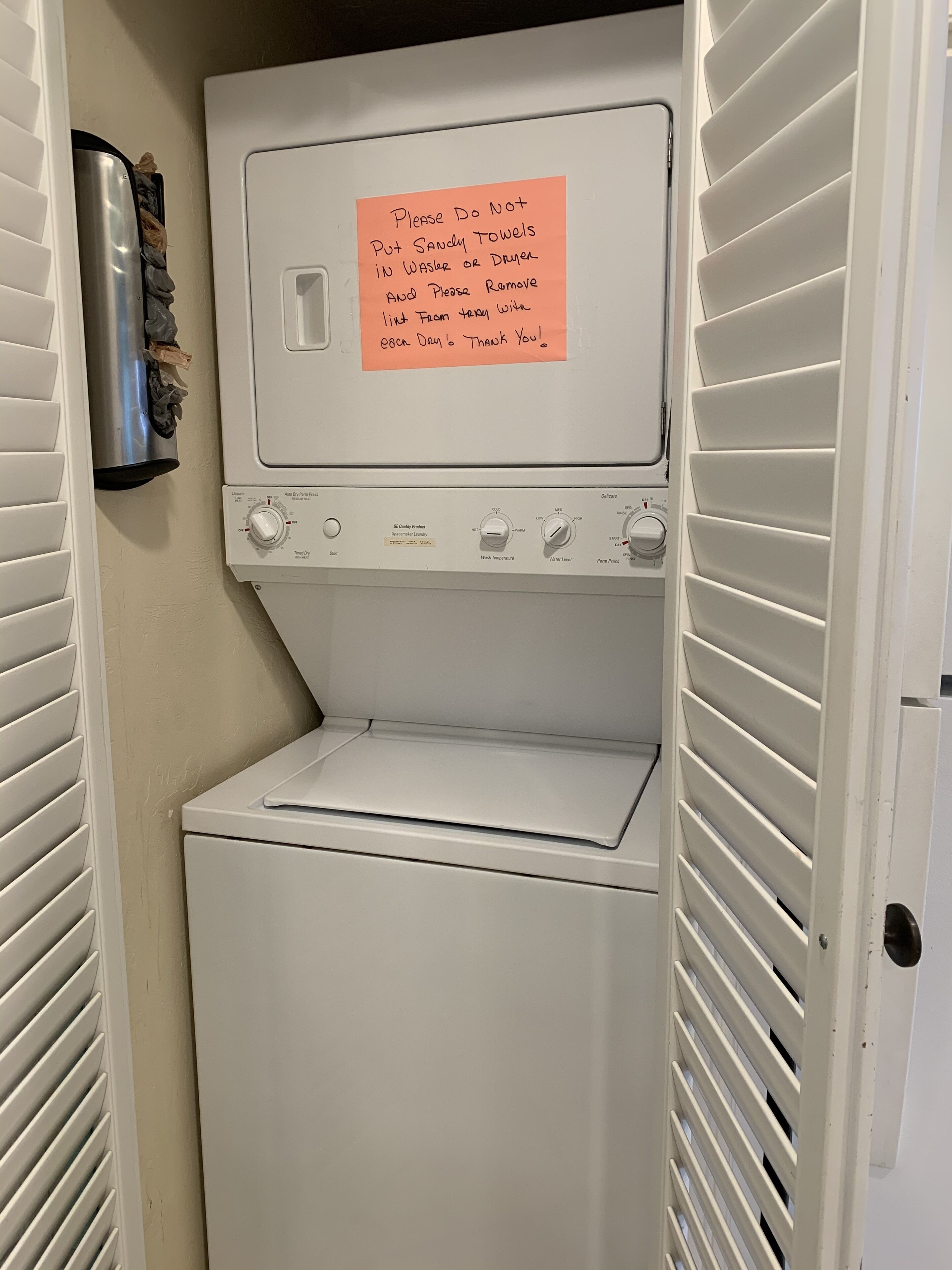 A front-loading washing machine with a note on the door requesting not to put sandy towels in the washer or dryer and to remove them daily.
