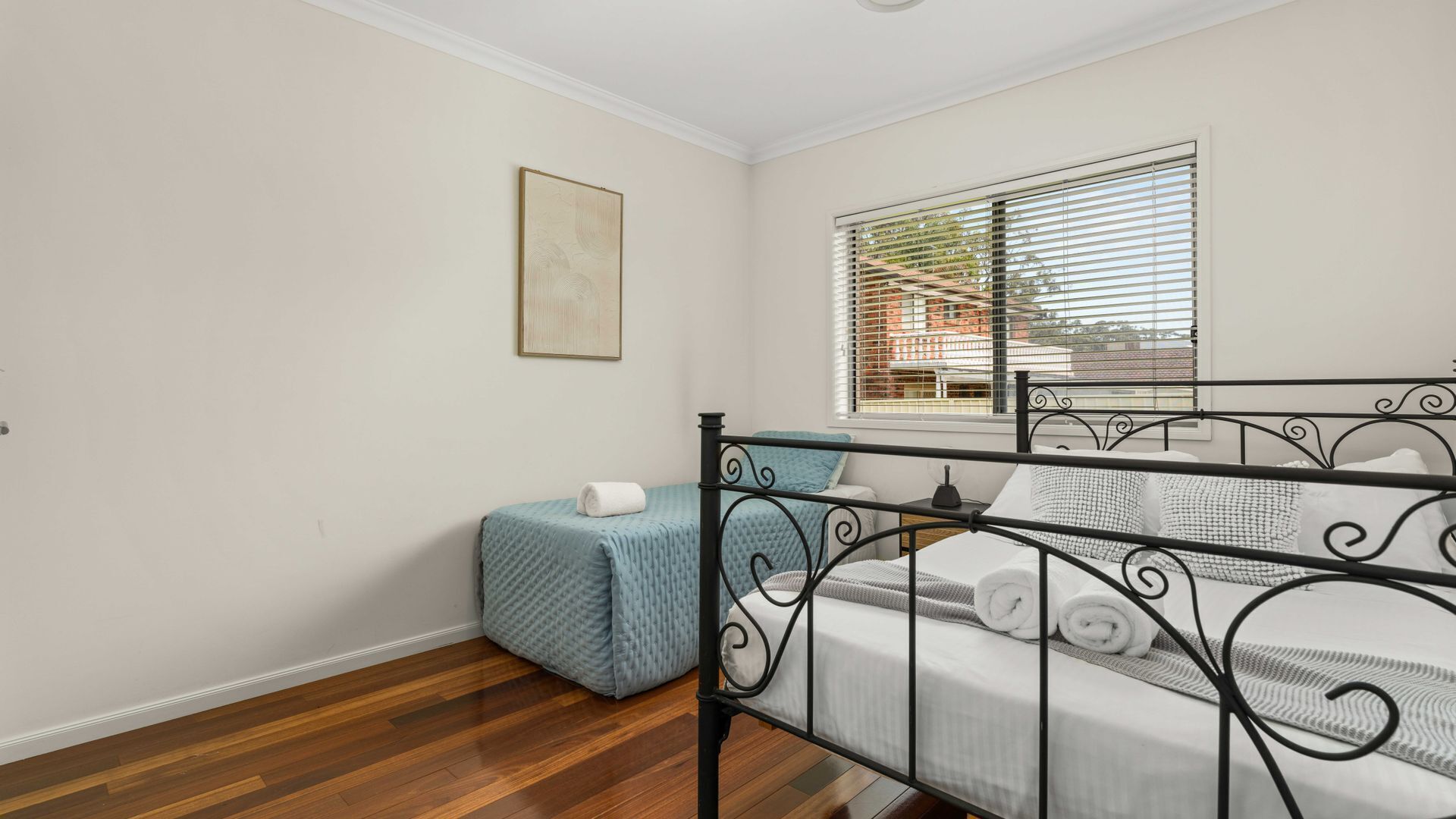 A bedroom with a bed, a window with blinds, a small bedside table, and a painting on the wall.