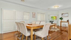 Uma sala de jantar com mesa e cadeiras de madeira, paredes brancas e janelas amplas com persianas.