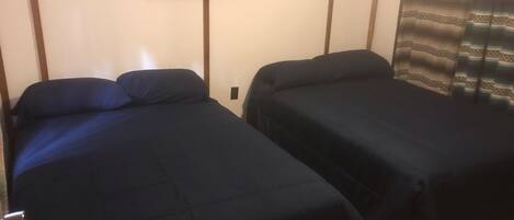 Dos camas individuales con ropa de cama azul oscuro en una habitación con pared lisa, un cuadro de paisaje enmarcado y una cortina estampada.