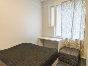 A small, simple room with a bed, a desk, and a window with blinds.