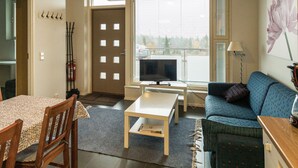 Ein Wohnzimmer mit einer Couch, einem Couchtisch, einem Fernseher und einem Esstisch mit Stühlen. Eine Tür mit Fenster und ein Feuerlöscher sind an der Wand angebracht.