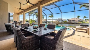A covered outdoor dining area with wicker furniture, a pool, and a view of palm trees and houses.