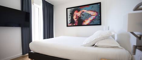 A modern hotel room with a large bed, a flat-screen TV mounted on the wall, and a painting above the bed.