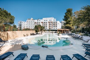 Un hotel con piscina rodeada de sillones y tumbonas, una terraza de madera y un edificio grande al fondo.