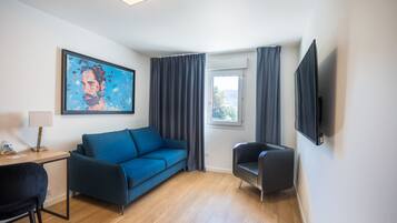 Una habitación moderna con un sofá azul, un sillón negro, un escritorio con lámpara, un cuadro en la pared, un televisor montado y una ventana con cortinas.