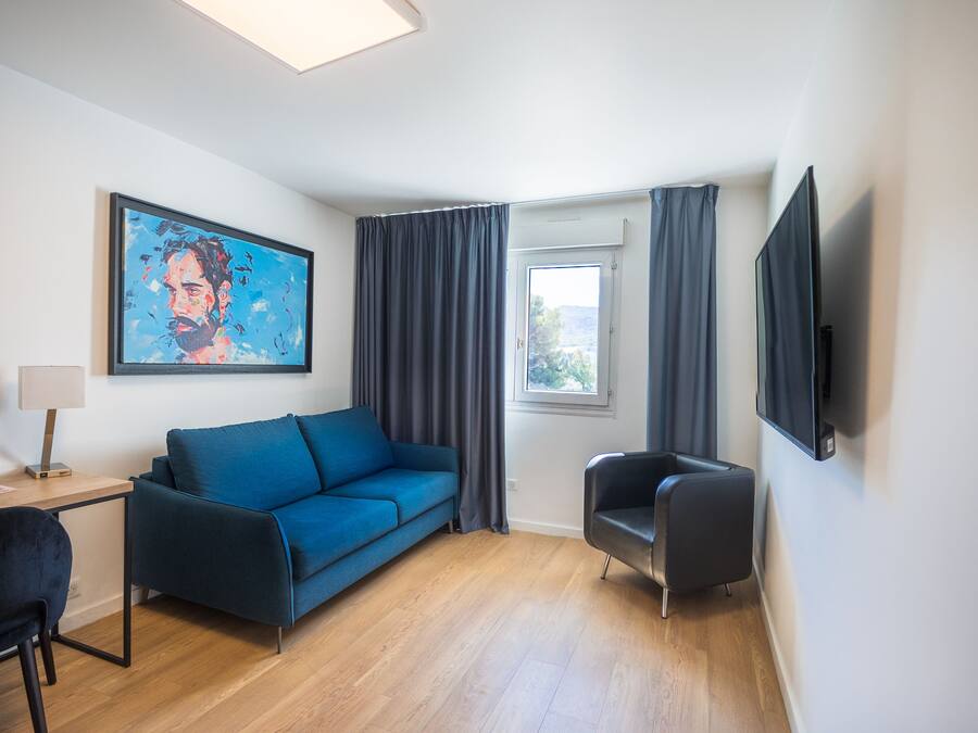 Una habitación moderna con un sofá azul, un sillón negro, un escritorio con lámpara, un cuadro en la pared, un televisor montado y una ventana con cortinas.