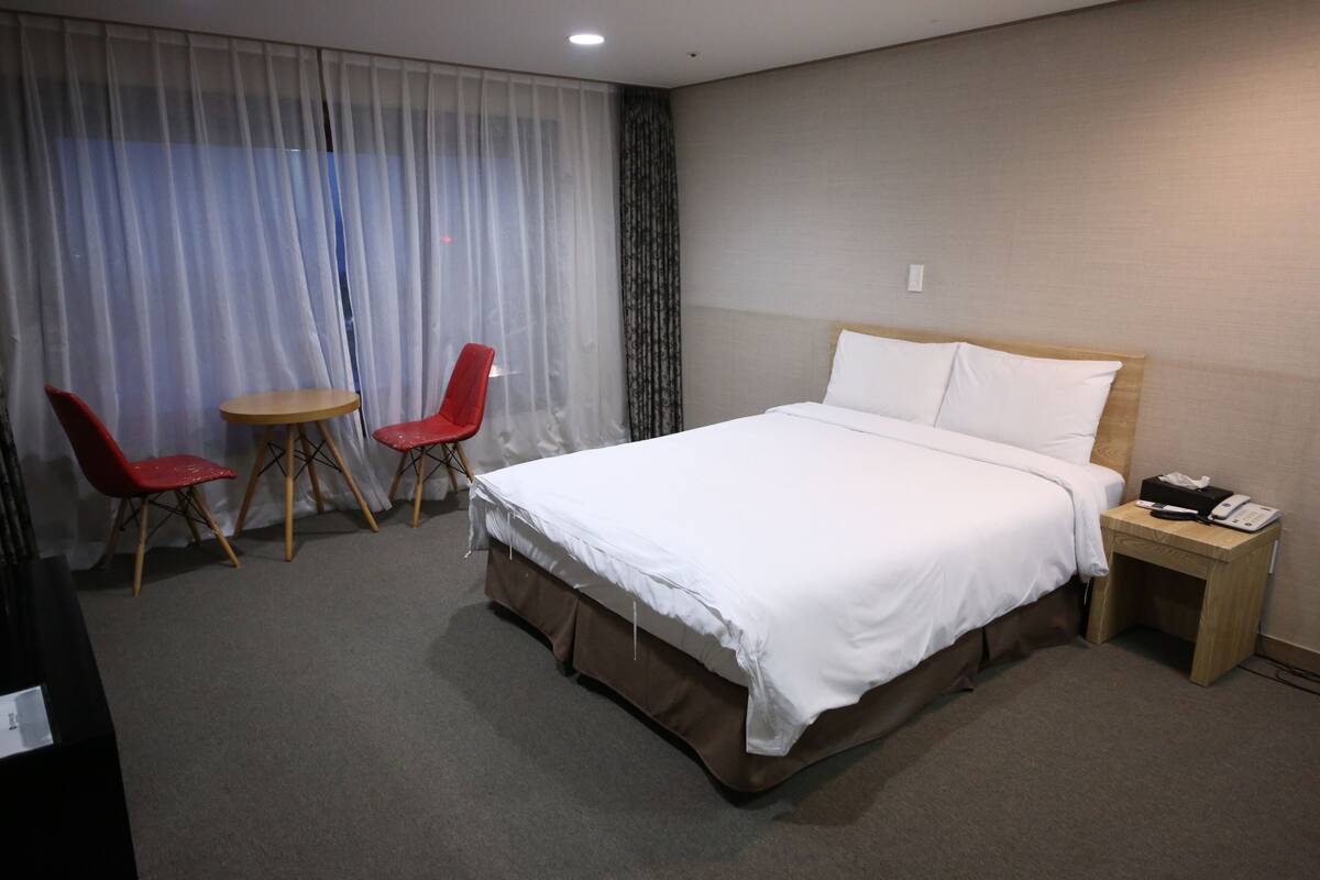 A hotel room with a large bed, a bedside table with a phone, two red chairs, a small round table, and sheer curtains.