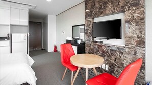 A hotel room with a bed, a round table, two red chairs, a TV mounted on a marble wall, a minibar, and a door leading to a bathroom.