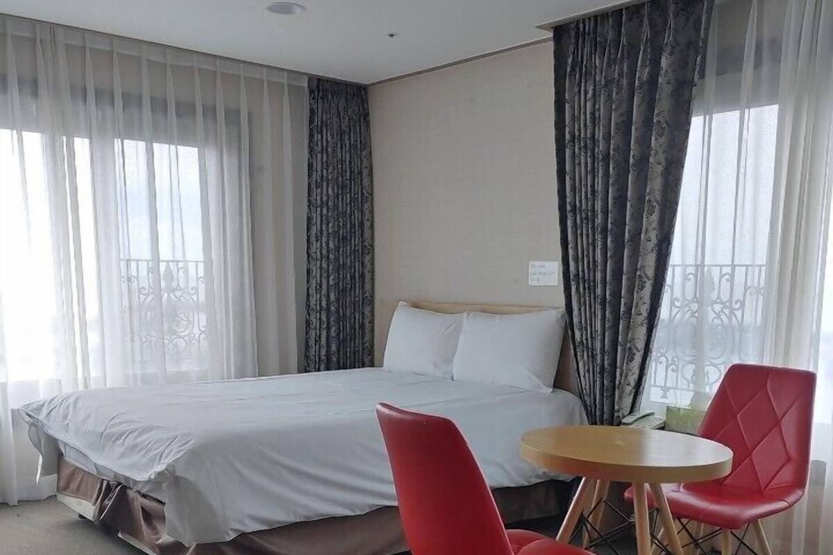 A hotel room with a bed, two red chairs, a small round table, and sheer curtains on the windows.