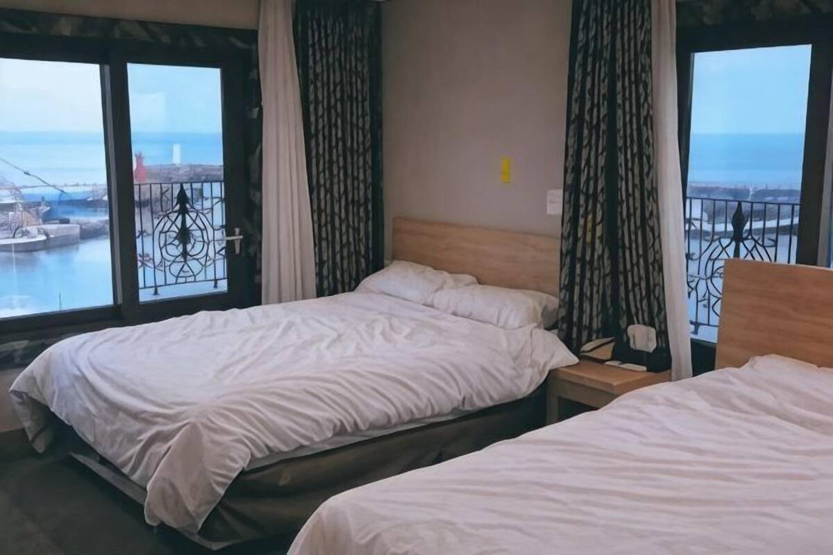 A hotel room with two beds, a wooden headboard, a nightstand, a window with a view of the sea, and a balcony with a railing.
