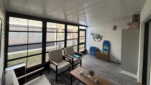 A sunlit room with large windows, a wooden bench, two chairs, and a small table with a plant and decorative items.