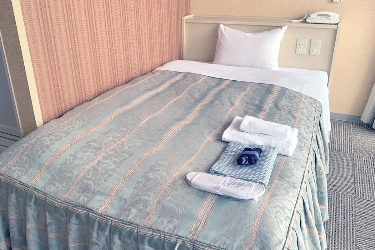 A hotel room with a bed, pillows, a folded towel, a phone, and a wall-mounted control panel.