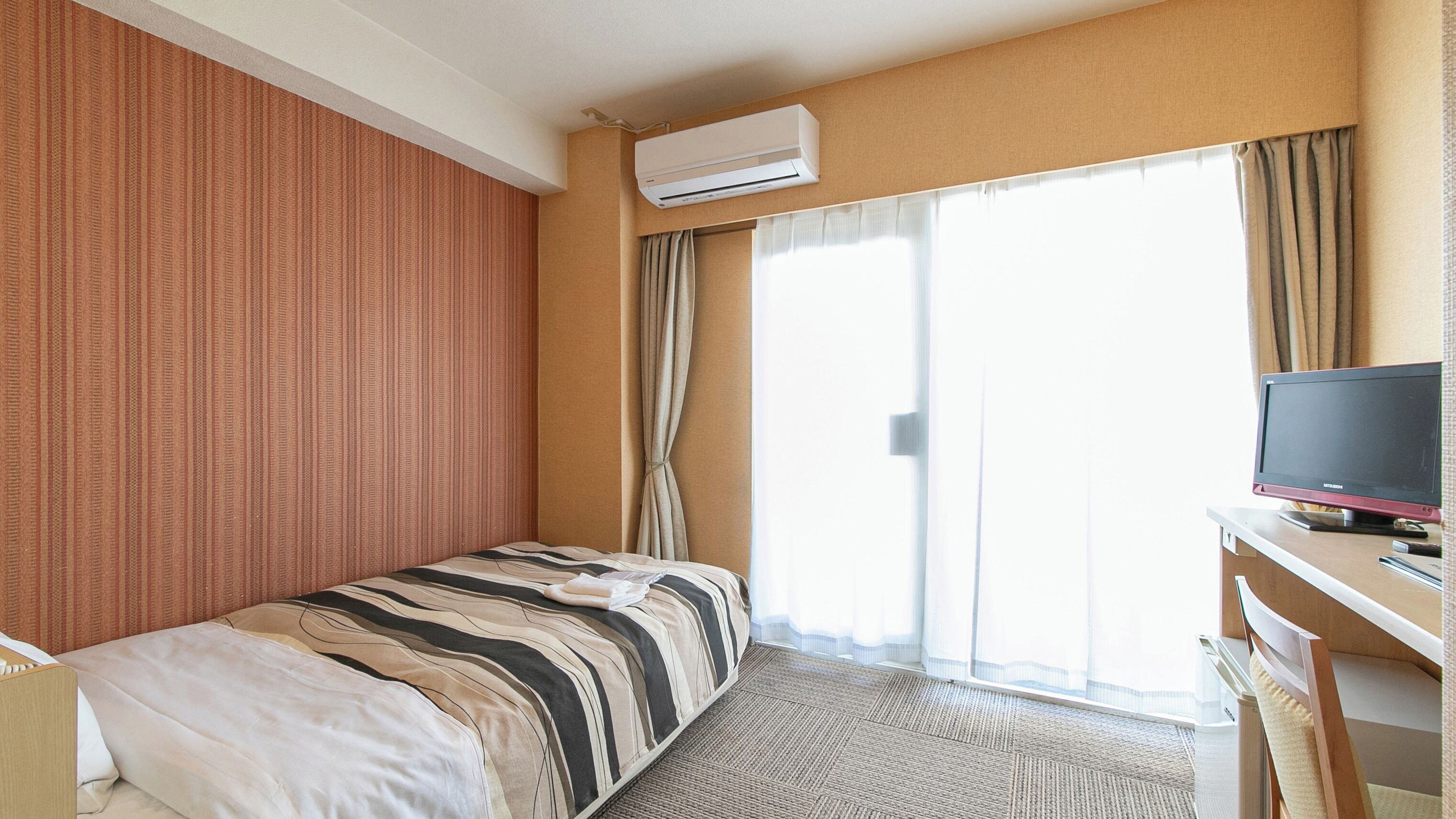 A hotel room with a bed, a desk with a computer, an air conditioner, and a large window with curtains.